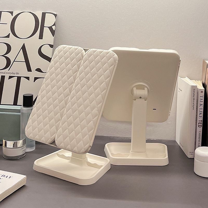 White quilted makeup mirror on a gray surface with books and a magazine in the background.