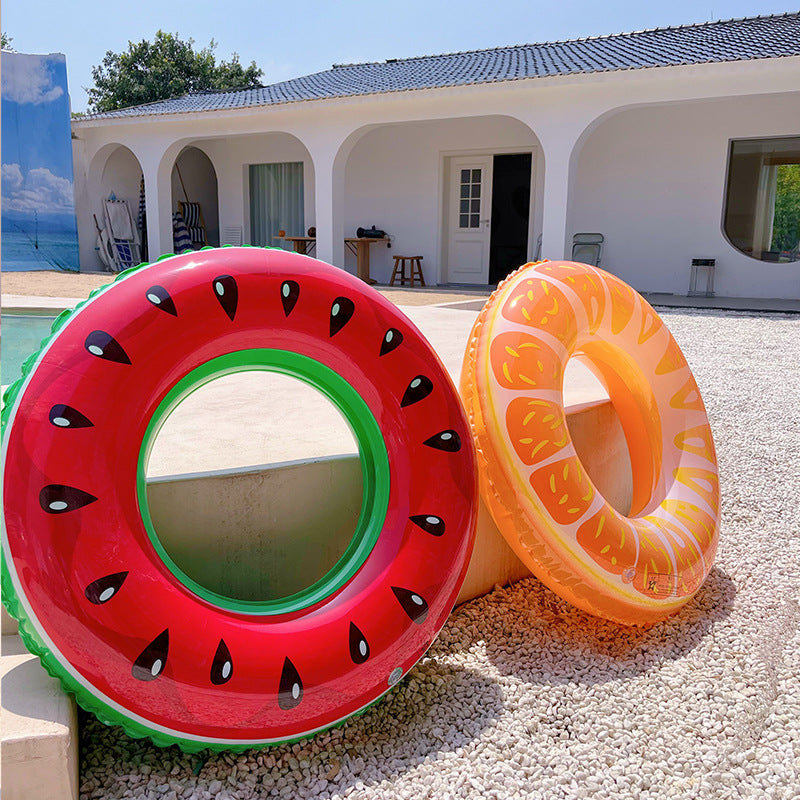 Two inflatable fruit-themed rings on a pebbled surface with a house in the background.