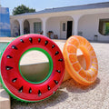 Two inflatable fruit-themed rings on a pebbled surface with a house in the background.
